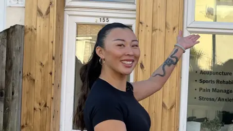 Rebecca Chung smiles outside her business. She has long black hair that is tied back behind her head. She wears a black t-shirt and gold hoop earrings. She holds one arm out toward her business and a snake tattoo can be seen on the inside of this arm.