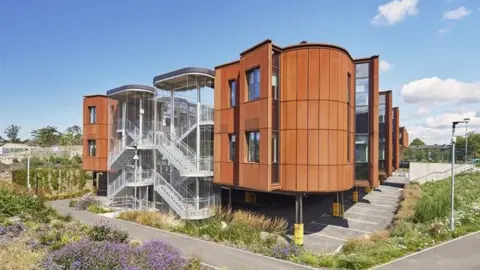 A modern brown building with exposed staircases surrounded by greenery and flowers.