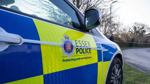 Essex Police An Essex Police branded police car pulled up on the side of the road. It is parked. Near the road there are trees and branches.