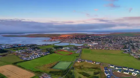 ORKNEY 2025 An aerial view of the Picky in Orkney. The sky is blue and grey. A number of green fields and houses are below. A running track surrounding a green sports pitch can be seen in the distance.