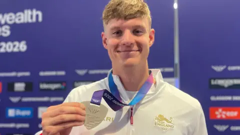 A man holding a silver medal with a purple ribbon around his neck. He is wearing a white Team England tracksuit top