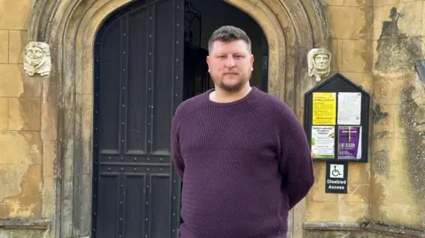 Eric Johnson/BBC Councillor Alistair Willoughby stands in front of the church door with his hands behind his back, wearing a purple knitted sweater. He has short styled hair and a neat beard and moustache. 