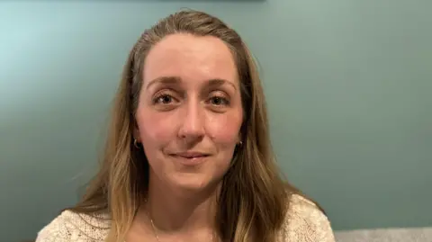 Celestina Mahovic has long light brown hair and is smiling. She is wearing a white jumper and is sitting in front of a teal wall.