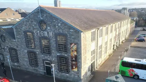 A large stone building with lots of windows with STEAM written on it, with parked coaches and cars outside