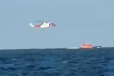 Diana Hare A helicopter hovers over an orange lifeboat on the water.