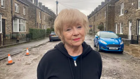 A woman with a blond bob hairstyle and black hooded top standing in the middle of terraced street