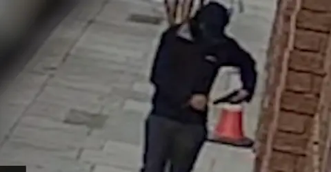 A hooded man dressed in black carries a gun in the street