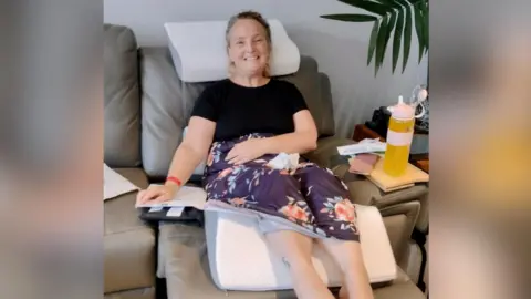 Family photo A woman in a black top and floral purple skirt is sitting on a grey sofa with her legs raised and a triangular white foam pillow under her knees.