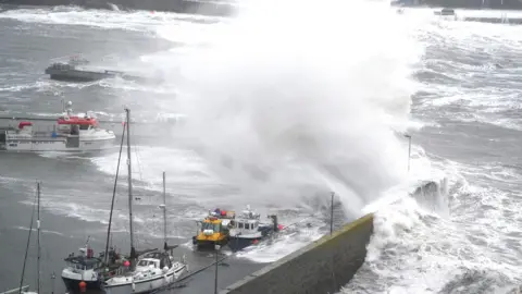 PA Media Large white waves crashing into a harbour over small boats that are tied up