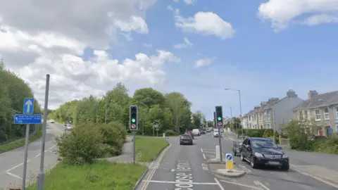A google street view image of Barncoose Terrace. There is a road going up to the left and a road straight ahead. There is green grass and a pedestrian footpath in the middle of the two. There are cars on the road driving in both directions. There are traffic lights which are green. There are houses on the right. 