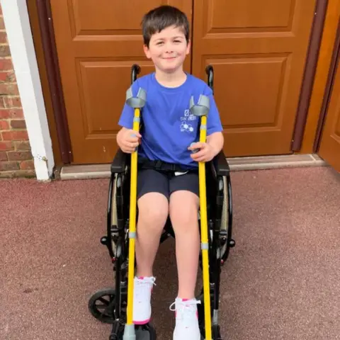 Family handout/PA Real Life Harry is seen in a wheelchair smiling while holding a pair of yellow crutches. 