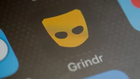 Getty Images A close-up of a mobile phone showing the Grindr app logo against a black background. The logo is a yellow mask with holes for eyes but none for mouth or nose, and it sits on a black background. The word Grindr appears beneath it in white text.
