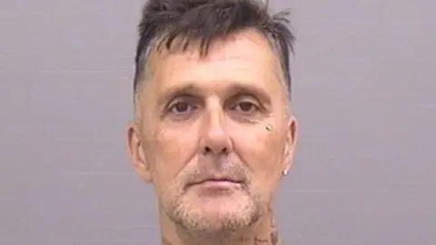 Custody photo of Simon Evans. He has stubble and very short cropped black hair.