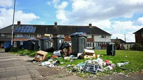 Getty Images Waste spills from overflowing and uncollected wheelie bins outside homes in the Shard End suburb of Birmingham, on 14 April 2025. The rubbish strewn over an area of grass includes carboard boxes, a cutlery tray, household waste in bags and litter.
