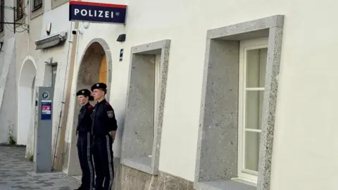 Two police officers stand outside a white-washed building with a police sign in German on the wall