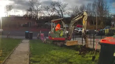 A workman in a high-visibility jacket is operating a digger. The digger is on a long, narrow strip of grass in a front garden leading from a house to a pavement. The machine is digging into the grass on the edge of the garden, up against a metal-bar fence separating the grass from a paved drive belonging to the house next door.