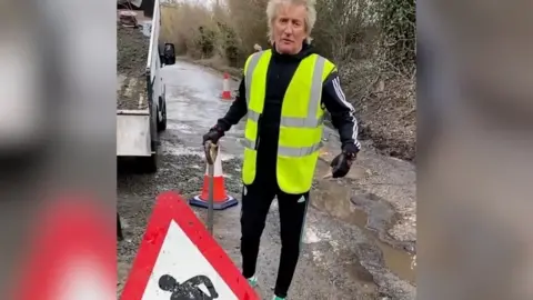 Rod Stewart/Instagram Sir Rod Stewart is standing on a road which has potholes filled with rain water. He is holding a shovel and is dressed in a black tracksuit and a yellow hi-vis jacket. Behind him is a lorry, emptying gravel onto the road