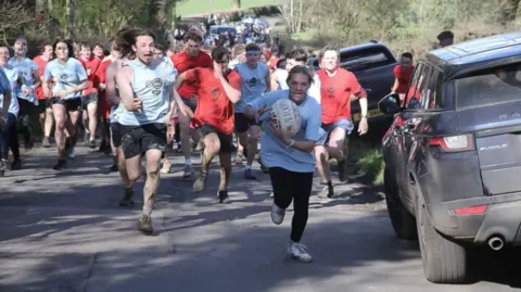 Chiddingstone Real Football A girl runs while carrying a ball in a white top. People in red and white tops chase her,