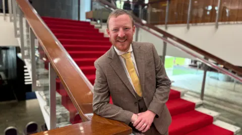 LDRS Liberal Democrat council leader Colin Ferguson, a man with fair hair and a short beard, wears a beige coloured suit, white shirt and yellow tie. He is smiling at the camera and is leaning on the wooden bannister of a large stair case with red carpet.
