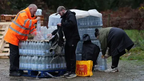 A person in an orange high-vis jacket handling a stack of bottled water in a car park with two members of the pubic loading bottles into bags.