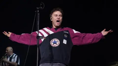 PA Media The singer Sam Fender stands with his arms open singing. He is wearing a purple NUFC top with the words Newcastle Brown Ale on it.