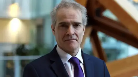 GOV.UK Charlie Taylor, Chief Inspector of Prisons, with white hair and a navy suit and purple tie. He looks impassively at the camera.