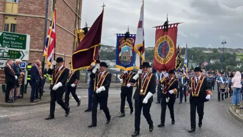 BBC A group of men wearing black suits and black bowler hats marching in a parade. They are wearing maroon sashes an carrying various flags. There is a band behind them.