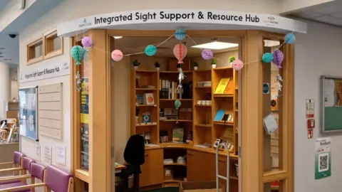 SIGHT MATTERS The hub situated inside Noble's Hospital. It is housed in a small, corner room fitted with wooden shelves, displaying various equipment. A sign above reads Integrated Sight Support and Resource Hub.