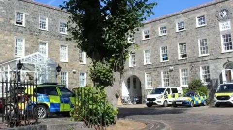 BBC Guernsey Police headquarters - two buildings in an L shape with grey and brown bricks, white panelled windows and four police cars parked outside, a large tree in the middle, on a sunny day 