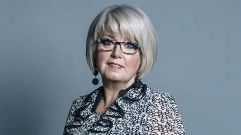 UK Parliament Baroness Helen Newlove, a woman with bobbed light blonde hair and glasees, wearing a black and white pattered blouse and she stands against a grey background. 