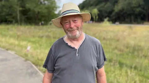 Archie Simpson Tarling/BBC A person wearing a wide-brimmed straw hat and a dark grey short-sleeved shirt stands on a paved path beside a grassy meadow. The background is softly blurred, showing tall grass and trees in a park-like setting. The photograph is a waist-up portrait taken outdoors in natural daylight.