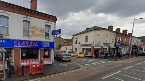 Google street view of a road junction. A fish and chip kebab house can be seen on the left, with a blue sign and a bus shelter outside. On the opposite corner is a south Asian sweet shop with a grey sign. Pedestrians can be seen walking on the pavement and other shops in the distance occupying the ground floors of terraced buildings.