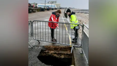 Borough Council of King's Lynn & West Norfolk Two women looking through a silver coloured fence at a sinkhole on the edge of a seaside promenade at Hunstanton, Norfolk. One is pointing down the hole. The promenade stretches out behind them. The beach is on the right of the frame.