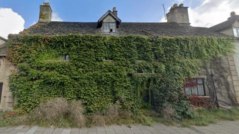 West End House, pictured in September 2023, is covered with leaves and growth, which covers windows and a door. Weeds and grass covers the base of the building 