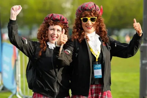 Elaine Livingstone Two kiltwalkers wearing long ginger wigs, red tartan hats and kilts with big white ruffs. They are smiling, punching the air and giving a thumbs-up. One has yellow glasses on. 