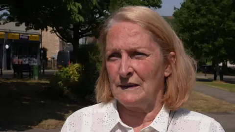 Casey Tremain talks to the camera. She has neck length thick blonde hair and is wearing a white shirt with spots on