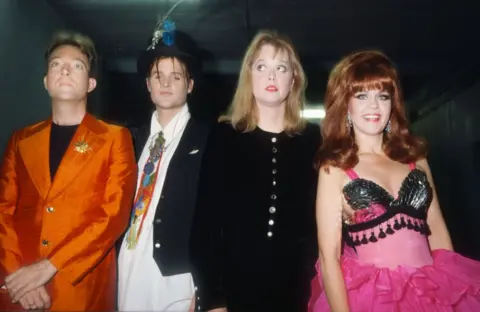 Getty Images The B-52s pictured backstage at a gig in 1989. Fred Schneider is wearing a striking orange jacket with a gold brooch, while Keith Strickland sports a feathered top hat. Cindy Wilson, in a relatively sober black outfit, glances to the side, while Kate Pierson, in her trademark beehive, is wearing a frilly pink tutu.