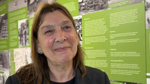 Julia Coutanche smiles slightly off-camera. She is wearing all black and has dark brown shoulder length hair. In the background is the museum exhibition.