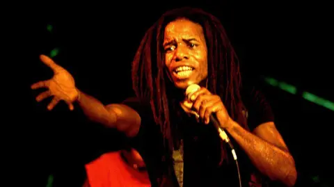 Getty Images: A young Eddy Grant with dreadlocks on stage, extending his hand to the audience while singing. 