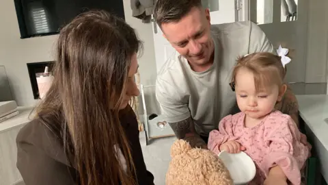 A couple in a grey living room and a little girl in a pink dress playing with a teddy bear.