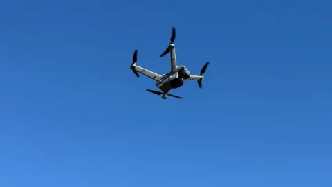 A drone in the air. The drone is grey and black with four arms with rotors on the end of them. It is against a purely blue sky, in the air.