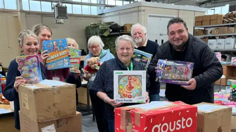 A group of charity volunteers along with BBC Radio Suffolk presenter Wayne Bavin smile at the camera in a warehouse. They hold children's toys which are being packed into cardboard boxes around them.