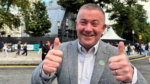 A man with grey/black hair smiling and giving thumbs up to the camera, he has a grey suit on