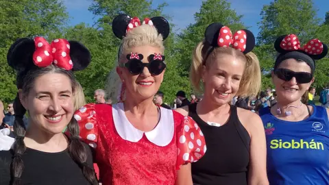 Four women standing next to each other. From left to right the first has plaited long brown hair, black top and is wearing Minnie Mouse ears. Next to her is Elaine who has blonde hair tied back wearing a red Minnie Mouse costume. Next to her is a lady with black clothing and short blonde hair wearing Minnie Mouse ears, and beside her is a lady with a blue top, red shorts black sunglasses and Minnie Mouse ears with her dark hair tied up.