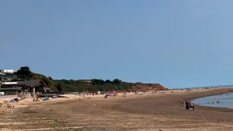 People enjoy a sandy beach at low tide, with swimmers in calm water and children playing near the shoreline under a wide blue sky.