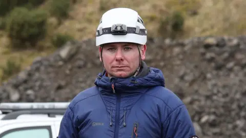 A man wearing a white helmet and a coat standing outside.