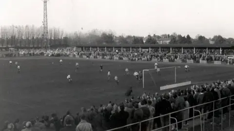 Eric Fernandez A black and white image of a football match taking place at Cambridge City's Milton Road ground. Men are scattered across the pitch and crowds of people can be seen standing and watching the match along the bottom and top of the ground. The people along the top of the ground are part-covered by a roof