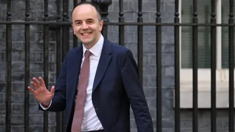 Britain's Health Secretary James Murray, who is wearing a suit and tie, waves to cameras while walking along Downing Street in London.