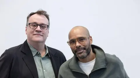 Max Cisotti/PA Wire Russell T Davies (left) wearing a black blazer, green shirt and black rimmed glasses and Benoit Swan Pouffer wearing a white t-shirt under a green fleece with a zip. They are both smiling.