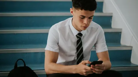 A teenage boy in school, wearing a shirt and tie, sat on some stairs, using his phone